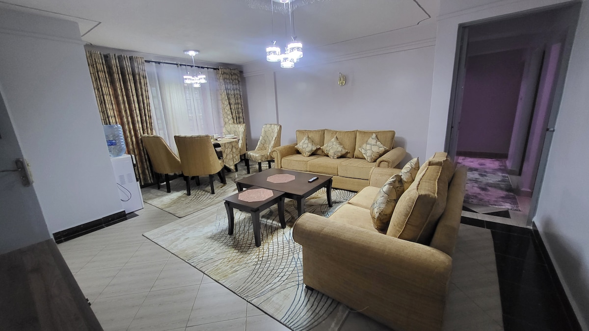 A spacious living area is furnished with a comfortable arrangement of beige couches and a dark coffee table. Elegant curtains drape over large windows, letting in natural light. A dining set occupies the corner, and decorative light fixtures add a touch of sophistication.
