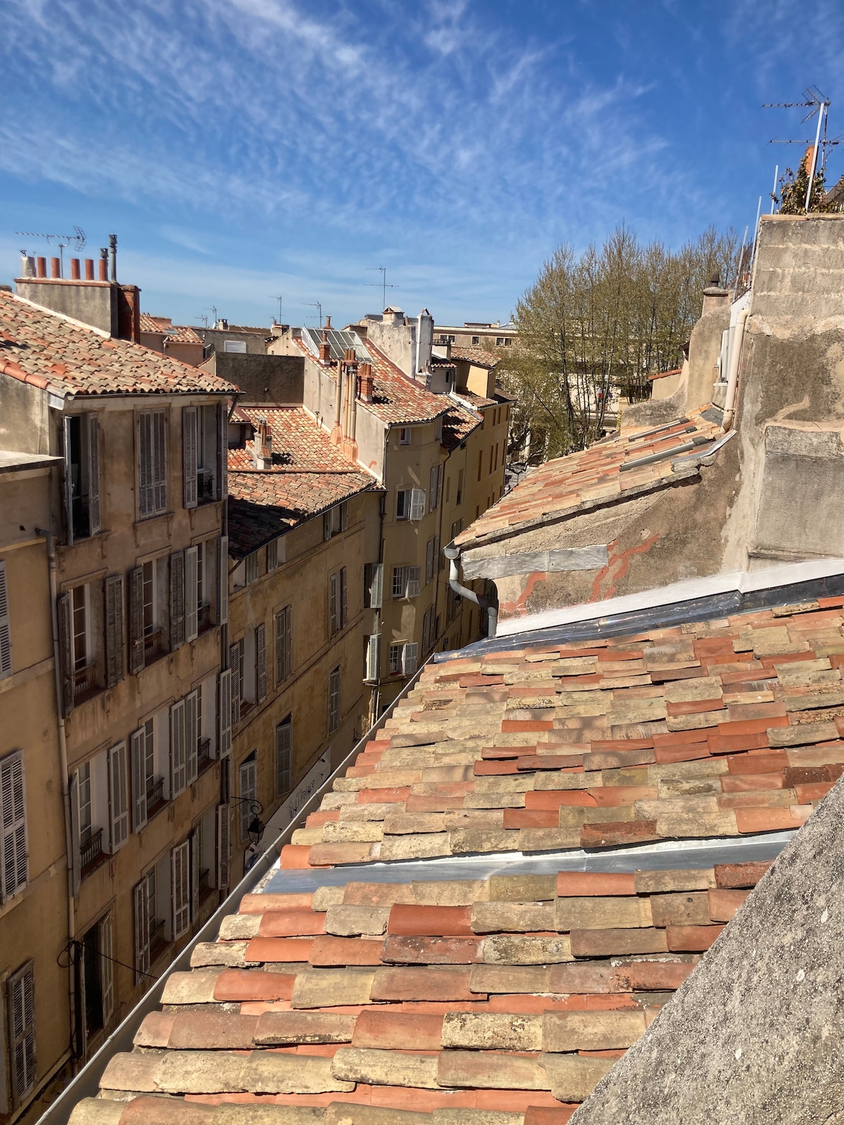 A view of the city rooftops features a patchwork of red and gray tiles. The adjacent buildings exhibit multiple windows and stone facades, with some greenery visible amid the structures. A clear blue sky with wispy clouds forms the backdrop.