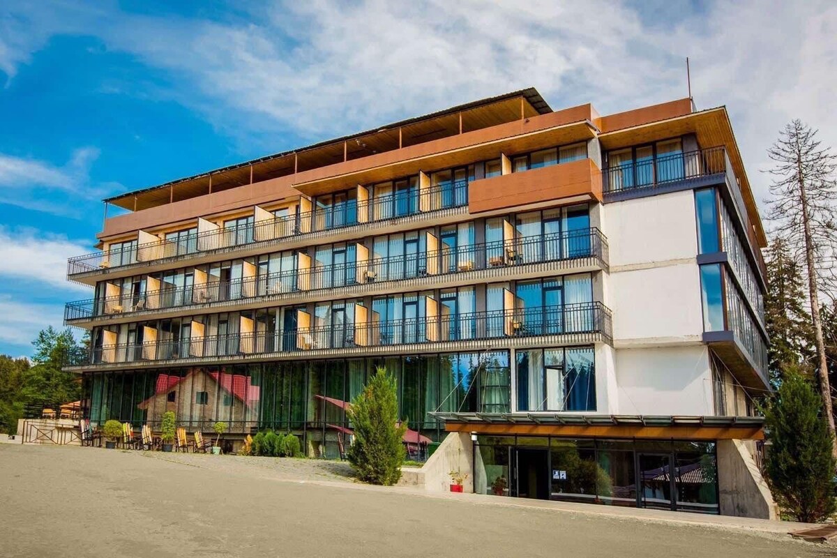 A modern hotel building with multiple levels is shown, featuring large glass windows and balconies that provide views of the surrounding forest. The structure is complemented by a clear blue sky and landscaped areas with greenery.