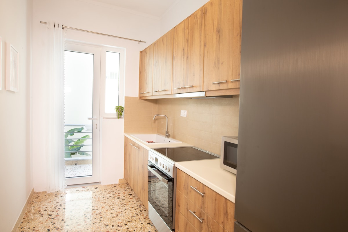 A modern kitchen is featured with wooden cabinetry and a light countertop. Stainless steel appliances are included, such as an oven and a microwave. A window provides natural light and offers a view of a small outdoor area with greenery.