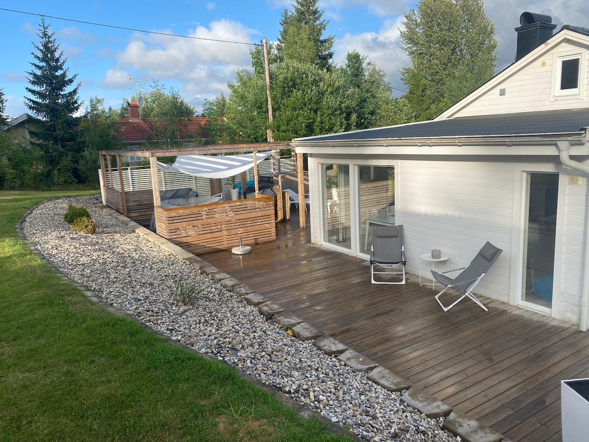 A spacious wooden deck is flanked by a pebble border and a lush green lawn. Two lounge chairs are positioned on one side, while a shaded seating area is visible, providing a relaxing outdoor space for guests. The surrounding trees offer a sense of privacy.