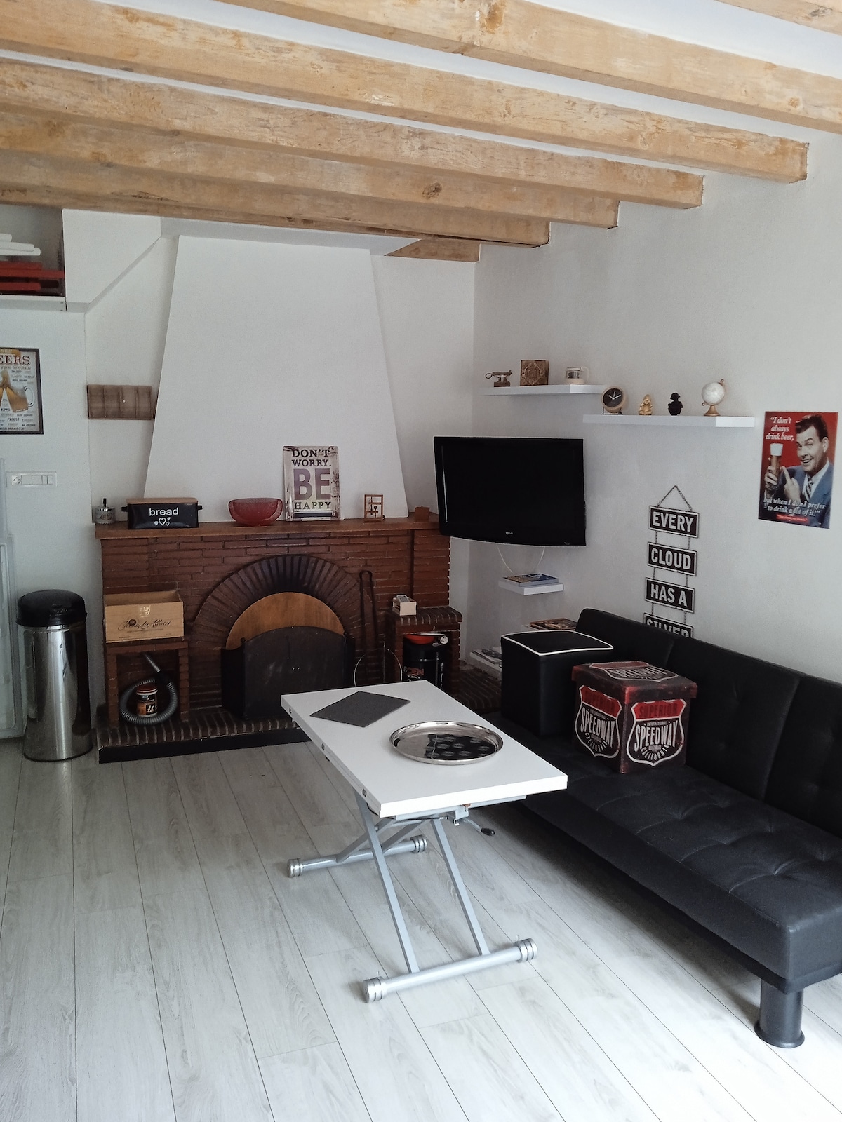 The living area features a black sofa facing a flat-screen television mounted on the wall. A coffee table stands in front of the sofa, and decorative shelves are positioned above the fireplace. Exposed wooden beams are visible on the ceiling, enhancing the space's character.