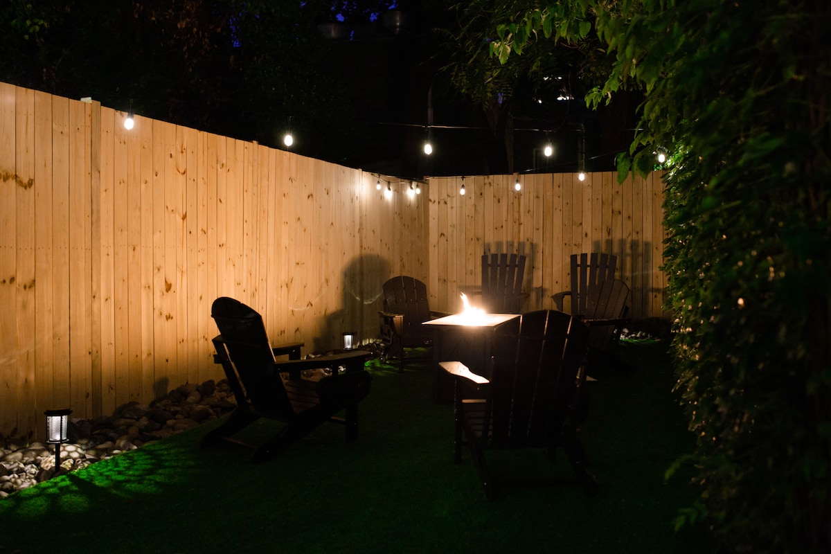 An outdoor space features a cozy arrangement of wooden chairs surrounding a fire pit, set against a wooden fence. Ambient lighting from string lights adds a warm glow, while green grass provides a soft surface for gatherings.