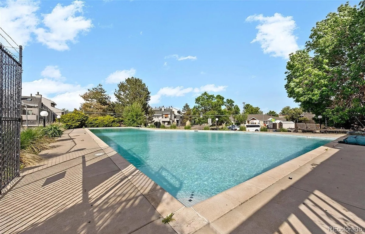A large outdoor swimming pool is shown, surrounded by greenery and residential buildings. The clear water reflects the sky, while lounge areas are positioned along the pool's edge. A fence encloses the pool area, providing a sense of privacy.