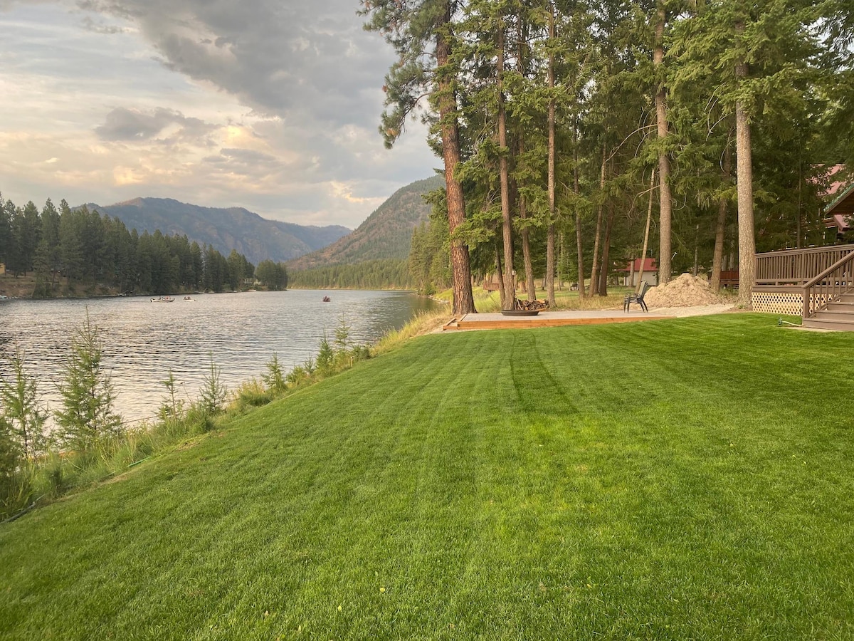 A green lawn gently slopes down to the Clark Fork River, framed by tall pine trees. The calm water reflects the surrounding mountains, while a fire pit area is visible near the water's edge. Soft light enhances the serene landscape.