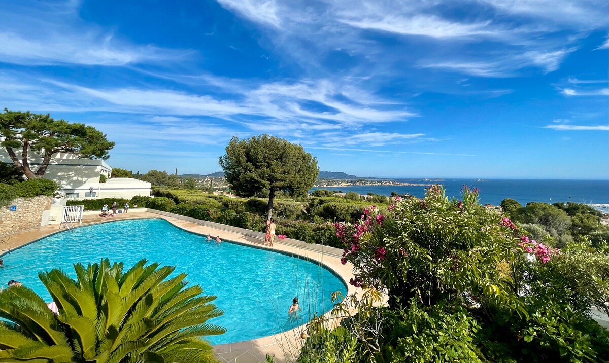 A spacious swimming pool is surrounded by lush gardens and vibrant flowering plants, providing a serene retreat. The panoramic view showcases the coastline and distant hills under a clear blue sky, inviting relaxation in a tranquil outdoor setting.