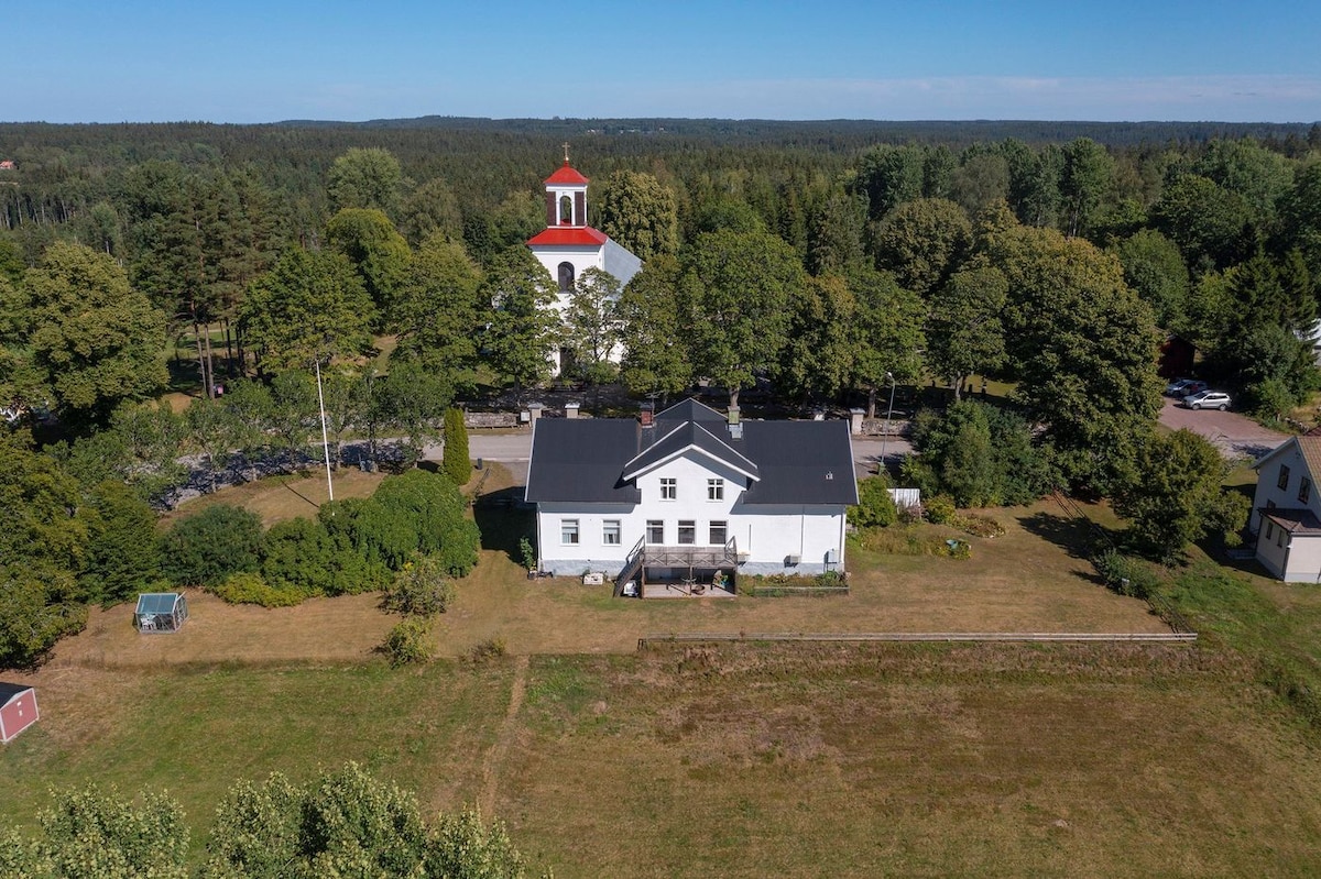 An aerial view showcases a large white building set amidst lush greenery, surrounded by trees. A tower with a red roof is visible in the background, indicating a nearby church. Open green fields extend in front of the property, enhancing the tranquil setting.