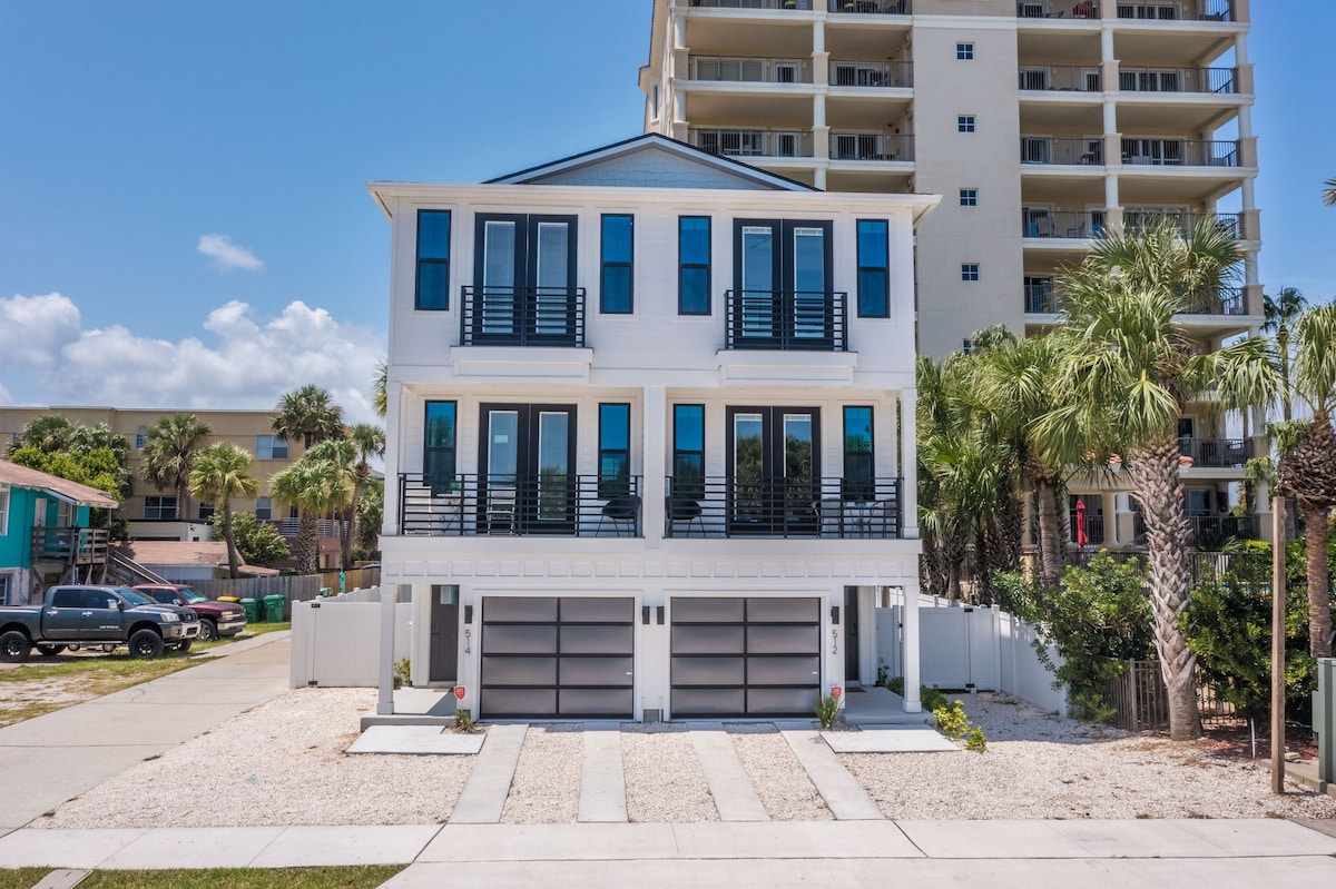 The exterior of the duplex showcases a modern three-story design with a light exterior, large glass windows, and two garage doors. A paved driveway is lined with pebbles, and palm trees add to the coastal vibe. The building stands prominently across from the beach.