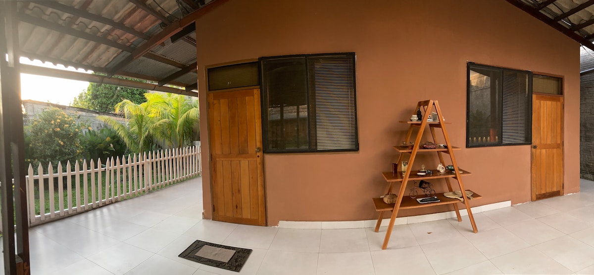 An exterior view of a building is presented, featuring a warm-toned wall and large windows allowing natural light. A wooden shelf is positioned against the wall, displaying various items. Lush greenery is visible in the background, alongside a white picket fence that frames the space.