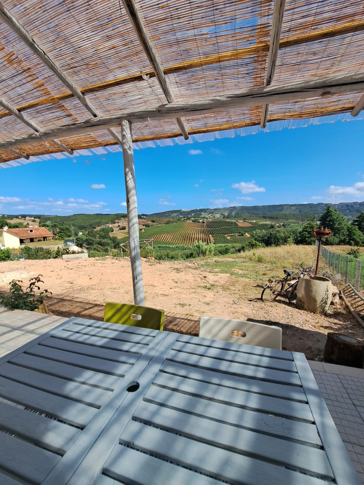 An outdoor dining area is visible, featuring a wooden table set against a backdrop of rolling vineyards. A shaded pergola offers respite from sunlight, while a panoramic view of hills and greenery stretches into the distance under a clear blue sky.