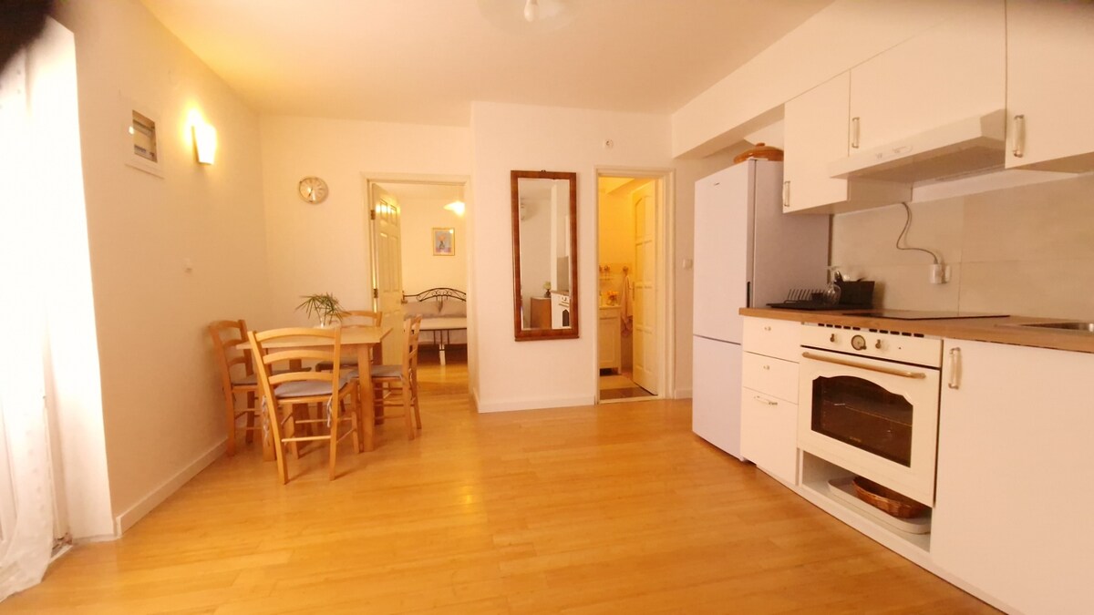 The light-filled kitchen and dining area feature wooden floors and white cabinetry. A dining table with four chairs sits invitingly, while a refrigerator and oven are clearly visible. An entryway leads to other spaces, enhancing the layout's openness.