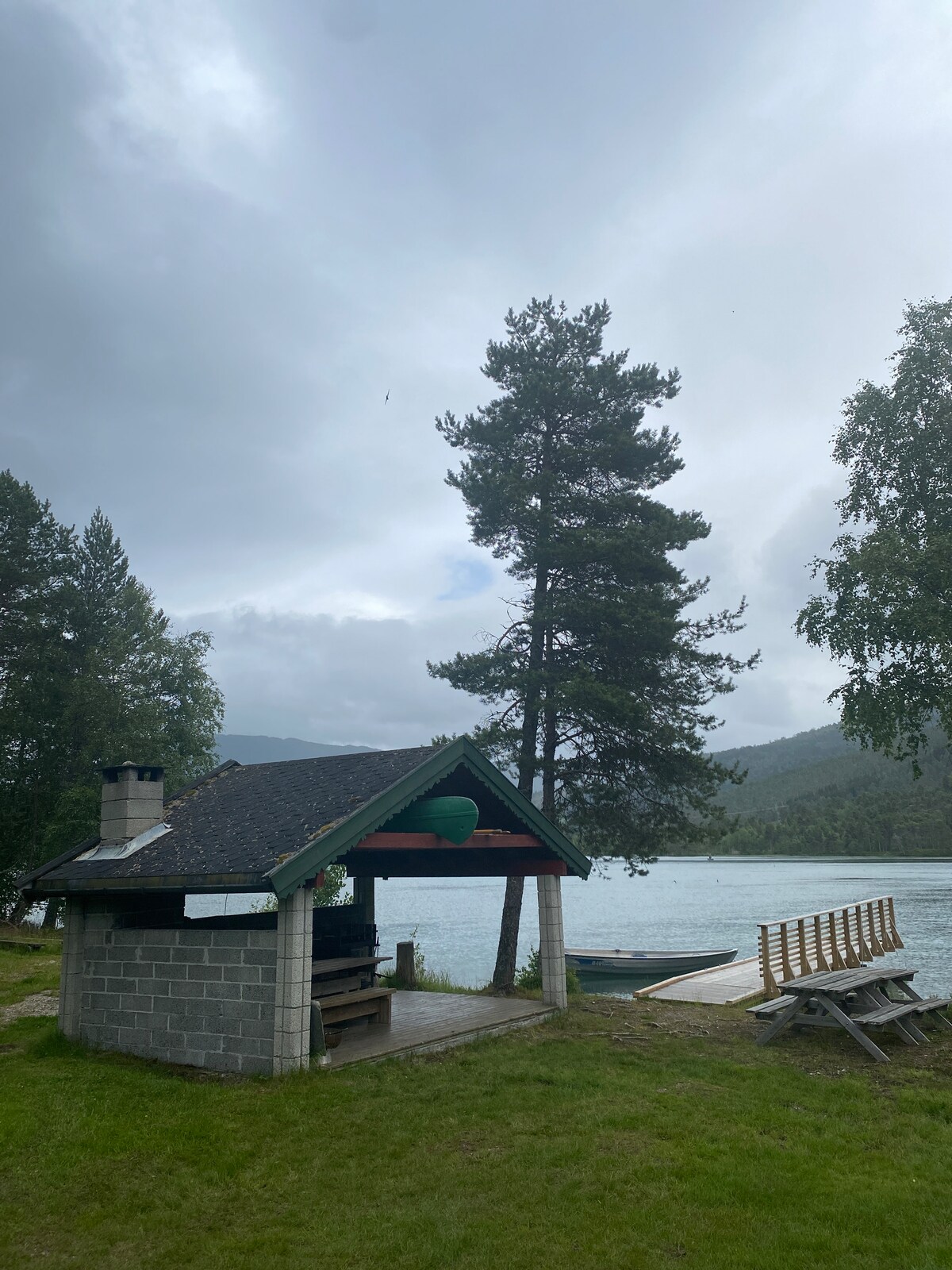 A sheltered grill cabin stands near a serene lake, surrounded by tall trees. A wooden bench is positioned in front, offering a view of the water and distant mountains. The overcast sky adds a calm ambiance to the tranquil setting.