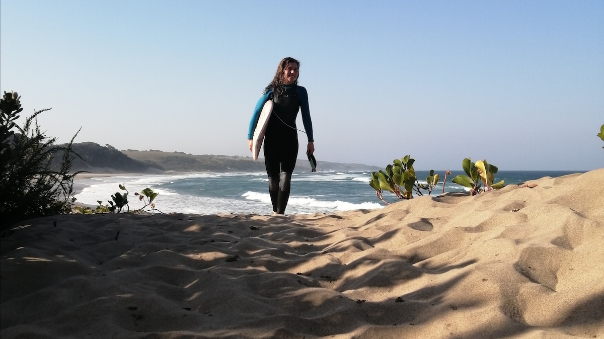 A person in a wetsuit is seen walking along a sandy path leading to the beach. The ocean waves can be observed in the background, with a clear blue sky above and greenery framing the scene.