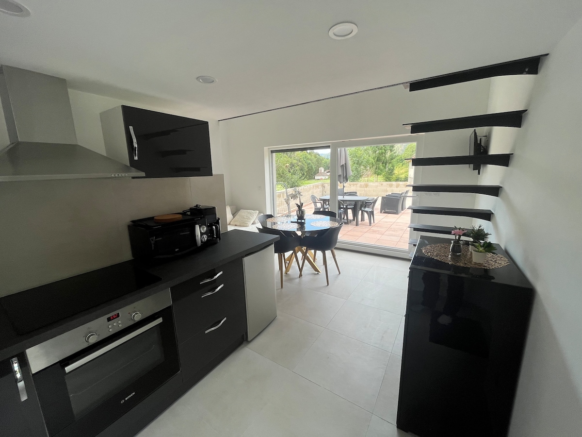A modern kitchen features sleek black cabinetry and appliances, with a cozy dining area nearby. A large window offers a view of the outdoor terrace and garden, allowing natural light to illuminate the space.