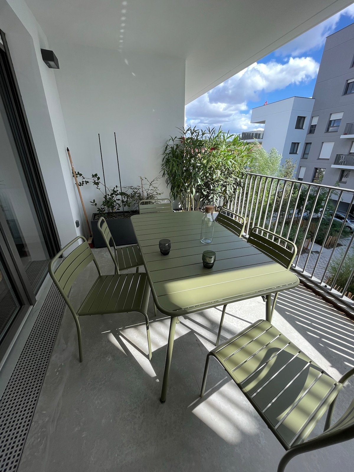 An outdoor terrace is furnished with a green metal dining table and matching chairs. Potted plants add greenery, and sunlight casts gentle shadows across the smooth concrete floor. The space offers a relaxed setting for meals or social gatherings.