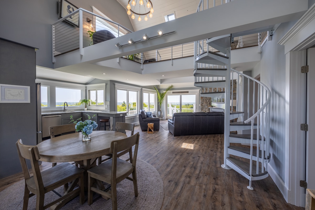An open floor plan is showcased, featuring a dining table that accommodates four. A modern spiral staircase leads to an upper level, while large windows offer sweeping views of Buzzard's Bay. Comfortable seating is arranged around the living area, enhancing the bright, airy atmosphere.
