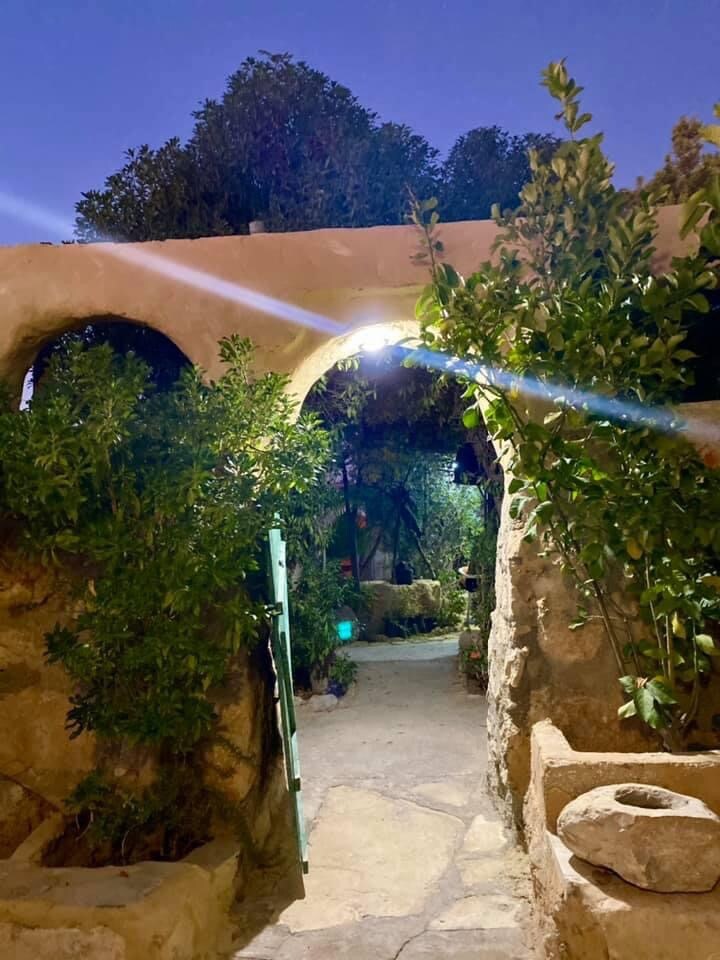 A stone pathway is framed by lush greenery, leading through an archway. Soft lighting highlights the entrance, creating an inviting atmosphere. The surrounding plants contribute to a serene environment, reflecting the rural charm of the area.