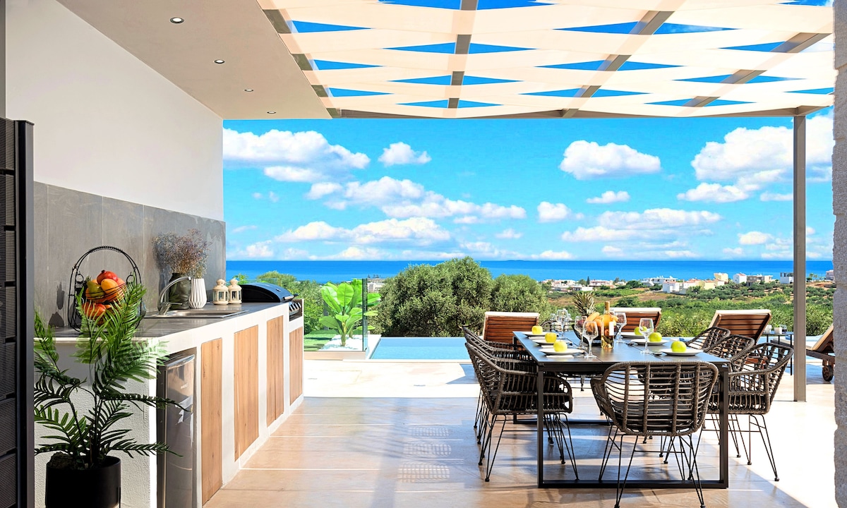 An outdoor dining area features a large table surrounded by stylish chairs, positioned under a lattice expanse. A well-equipped kitchen is visible, with ocean views framed by greenery and blue skies. The inviting space suggests an ideal setting for meals amidst a scenic landscape.