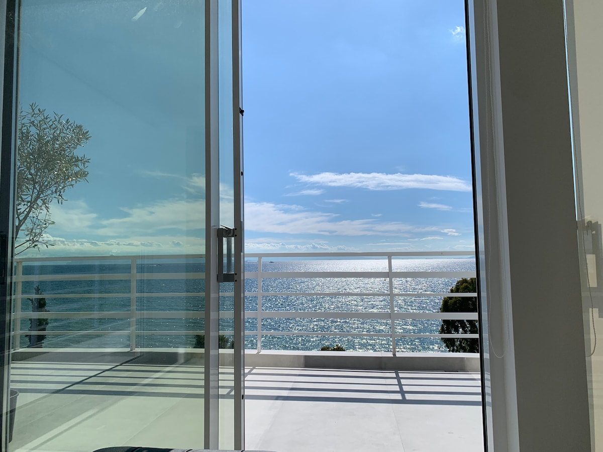 A view of the shimmering sea is framed by expansive glass doors, allowing natural light to fill the space. A spacious balcony with a white railing extends outward, inviting moments of relaxation while overlooking the tranquil waters.