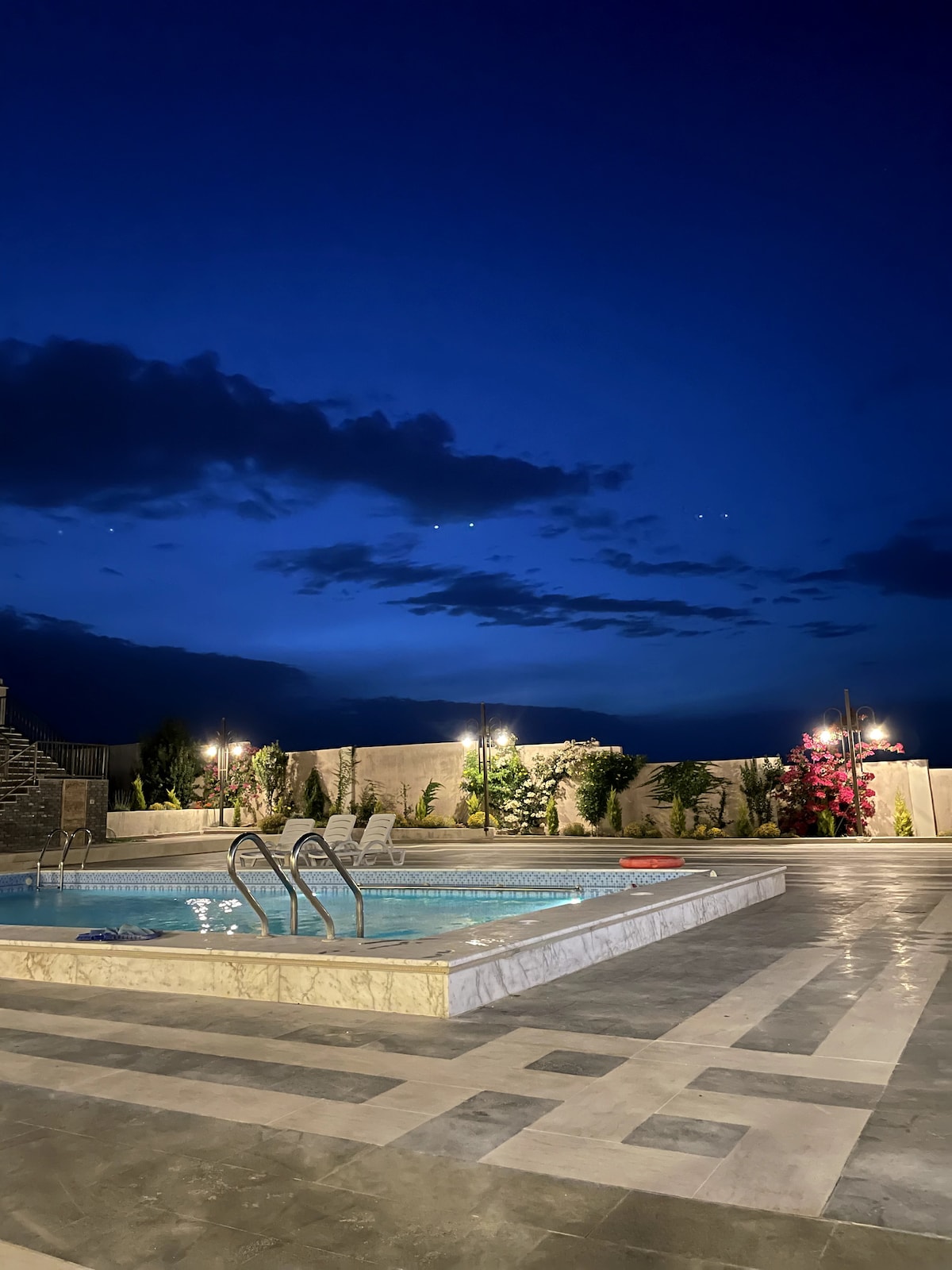 A large pool is illuminated under a deep blue sky, with clouds casting soft shadows. Surrounding greenery and colorful flowers add a touch of nature, complemented by soft lighting that highlights the pool's edges and the landscaped area.