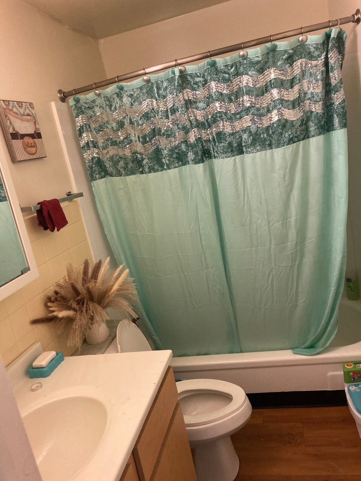 The bathroom features a light-colored countertop with a mirror above. A seafoam green shower curtain with decorative patterns hangs over the bathtub. A small plant adds a natural element to the space, with soft, neutral tones present in the decor.