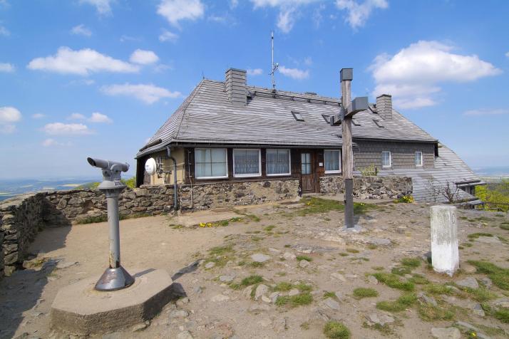 A charming mountain hut with a sloped wooden roof is situated on rocky terrain. The building features large windows and a welcoming entrance. Nearby, a stone observation point provides panoramic views, accompanied by a telescope for distant observations.