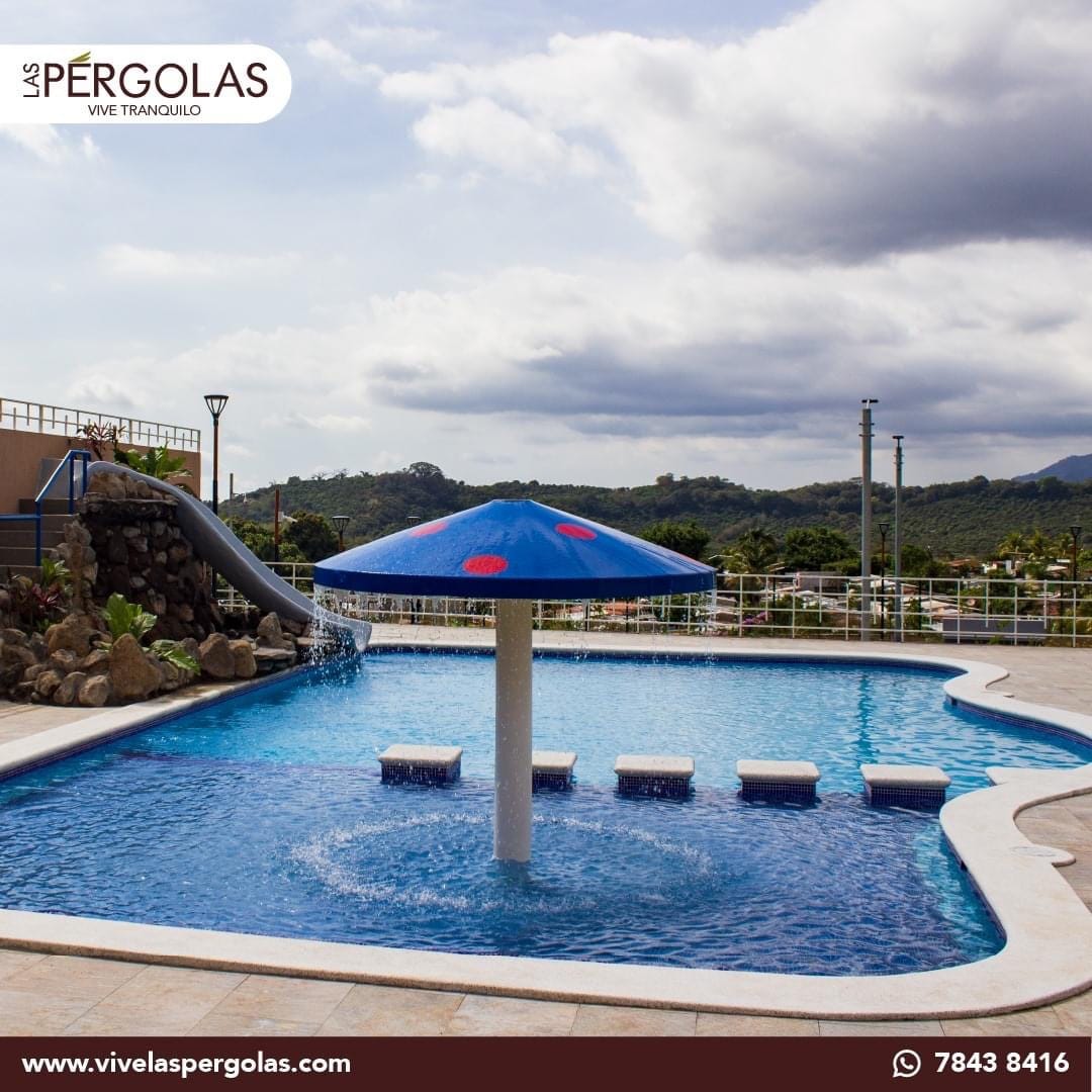 A large swimming pool features a distinct umbrella-shaped structure at its center, with water cascading down its sides. Several lounge chairs are positioned beside the pool, surrounded by a landscaped area and scenic views of the hills in the background.