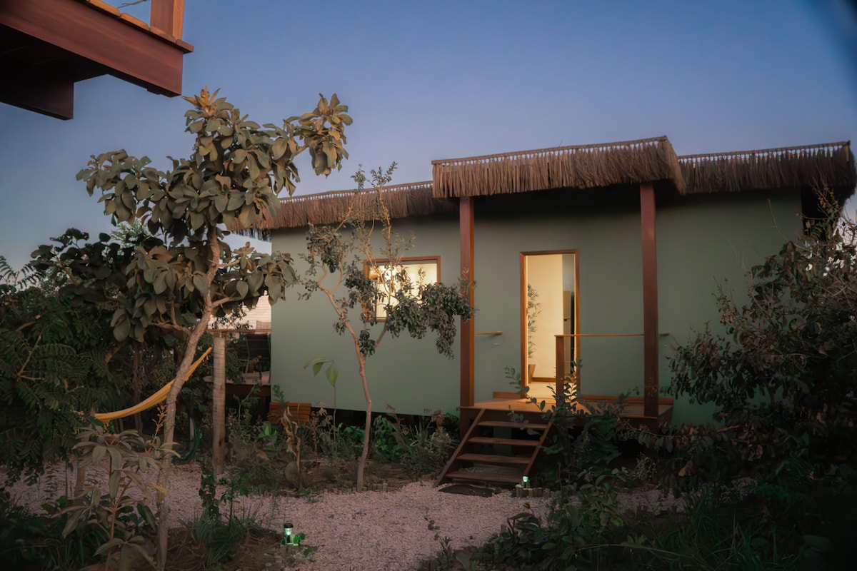 A serene small chalet is surrounded by lush greenery, featuring a welcoming entrance with wooden steps. Soft lighting from the windows hints at a cozy interior, while the thatched roof adds a natural touch to the overall design.