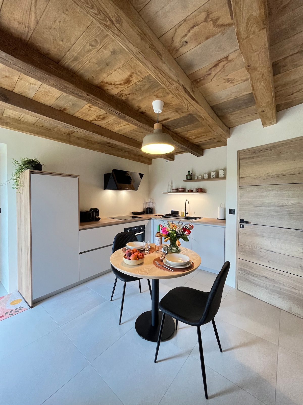 A modern kitchen is designed with wooden beams overhead, creating a warm atmosphere. A round table set for two, adorned with fruit and tableware, complements sleek cabinetry and essential appliances, ensuring a functional space for meal preparation and dining.