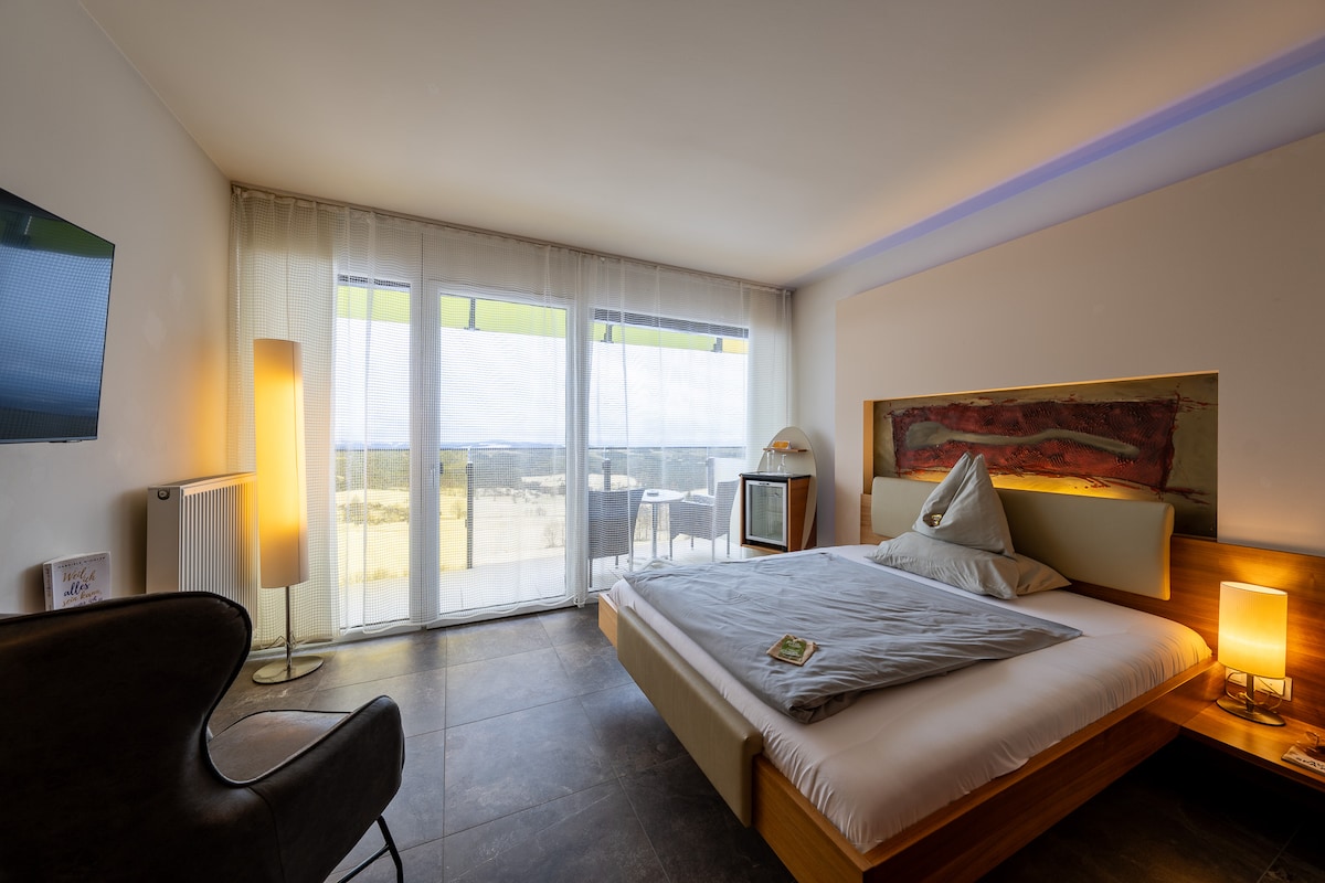 A contemporary bedroom features a spacious bed with light bedding and a decorative headboard. A comfortable chair is positioned nearby. Floor-to-ceiling windows allow natural light to fill the room and provide scenic views, while a sleek lamp adds a touch of warmth.
