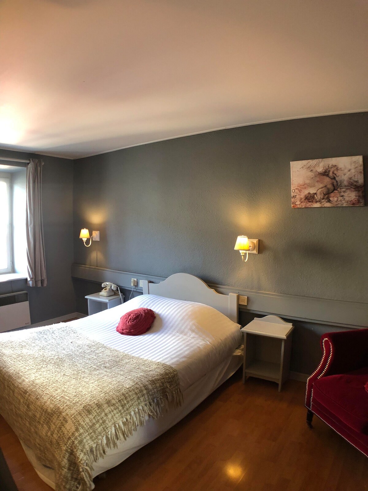 A cozy sleeping area is featured with a spacious bed dressed in soft white linens. A warm-toned throw blanket drapes over the bed, and a decorative pillow adds a touch of color. Soft wall sconces provide gentle lighting, while a small red armchair complements the neutral decor.