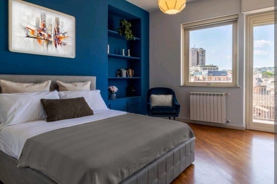 A spacious bedroom features a large bed with neutral bedding and a decorative pillow. Blue walls provide a calming backdrop, while a piece of abstract art hangs above. A comfortable chair is positioned nearby, and large windows allow natural light to flood the room.