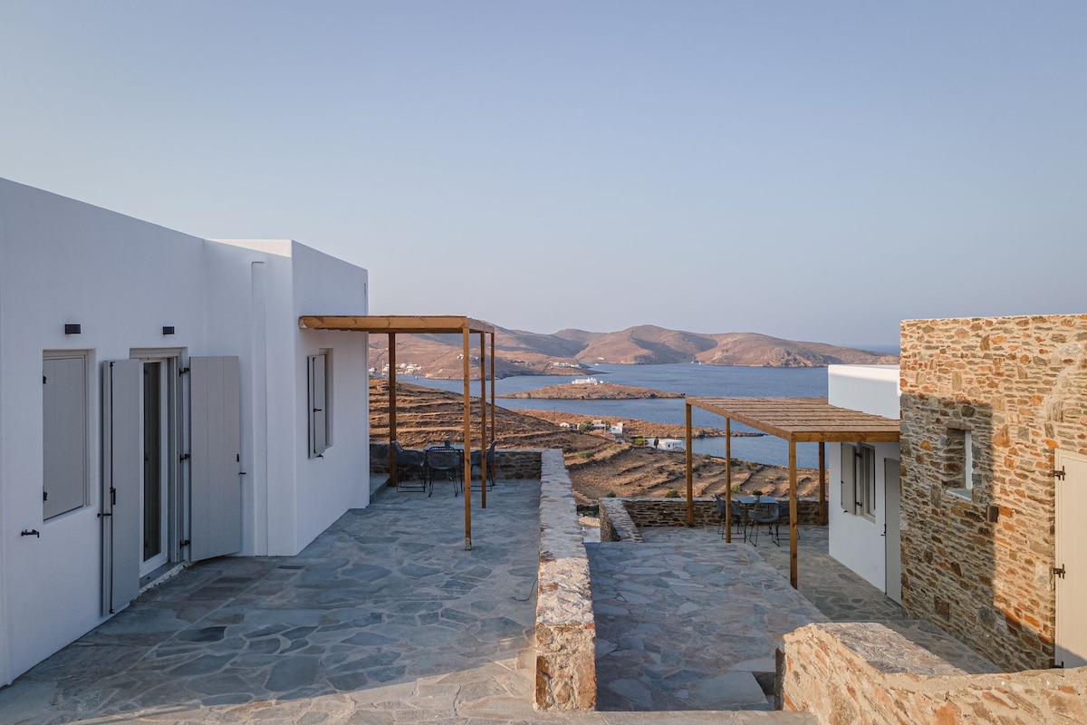 The outdoor area presents a stone pathway leading to light-colored buildings, accompanied by wooden pergolas providing shade. The backdrop features a sweeping view of the coastline and hills, with gentle waves lapping at the shore under a clear sky.