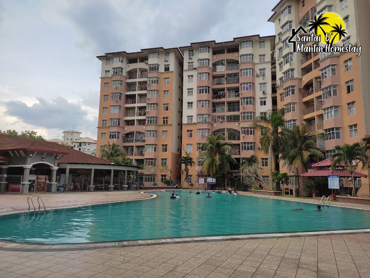 An outdoor swimming pool is surrounded by a spacious deck with palm trees. Adjacent to the pool, a multi-story building features balconies with glass doors. Water activities can be seen, with guests enjoying the pool area.