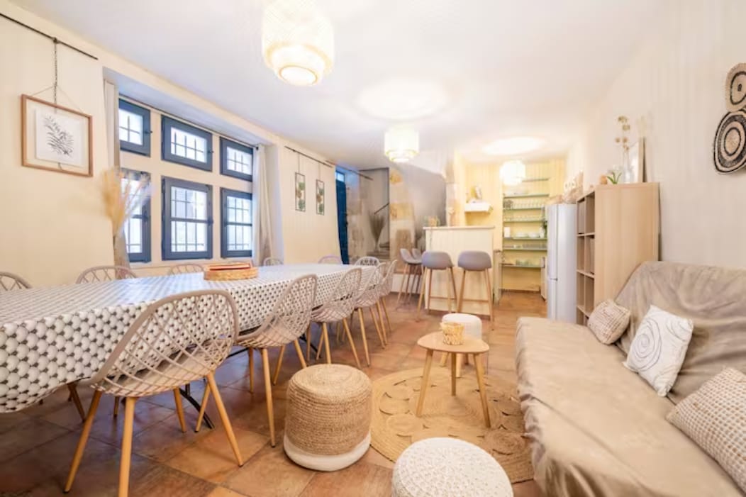 A spacious dining area features a long patterned table surrounded by rattan chairs. Soft lighting is provided by hanging fixtures, and a comfortable seating area is situated in one corner. Large windows allow natural light to fill the room, enhancing the cozy atmosphere.