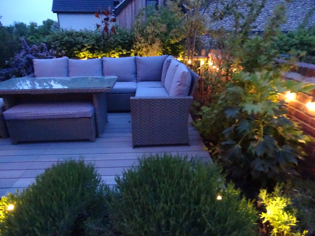 A landscaped garden area features a rattan-style corner sofa with two stools and a table, illuminated by solar lighting. Nearby, low shrubs and plants provide natural greenery, while the decking creates a cozy outdoor space, ideal for relaxation in the evening.