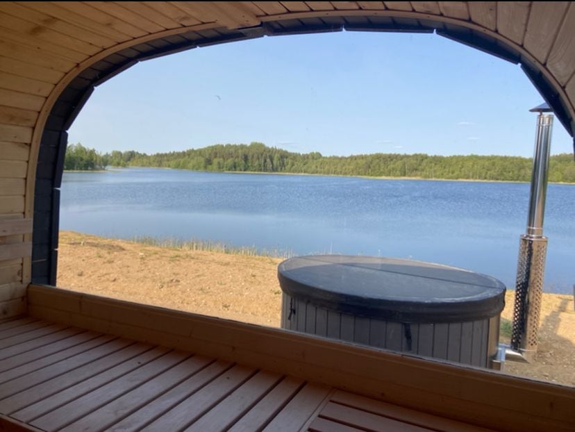 A wooden bench seat is positioned beneath a large arched window, offering a view of the calm lake and wooded shoreline. The scene features a round hot tub nearby, with natural elements enhancing the tranquil setting.