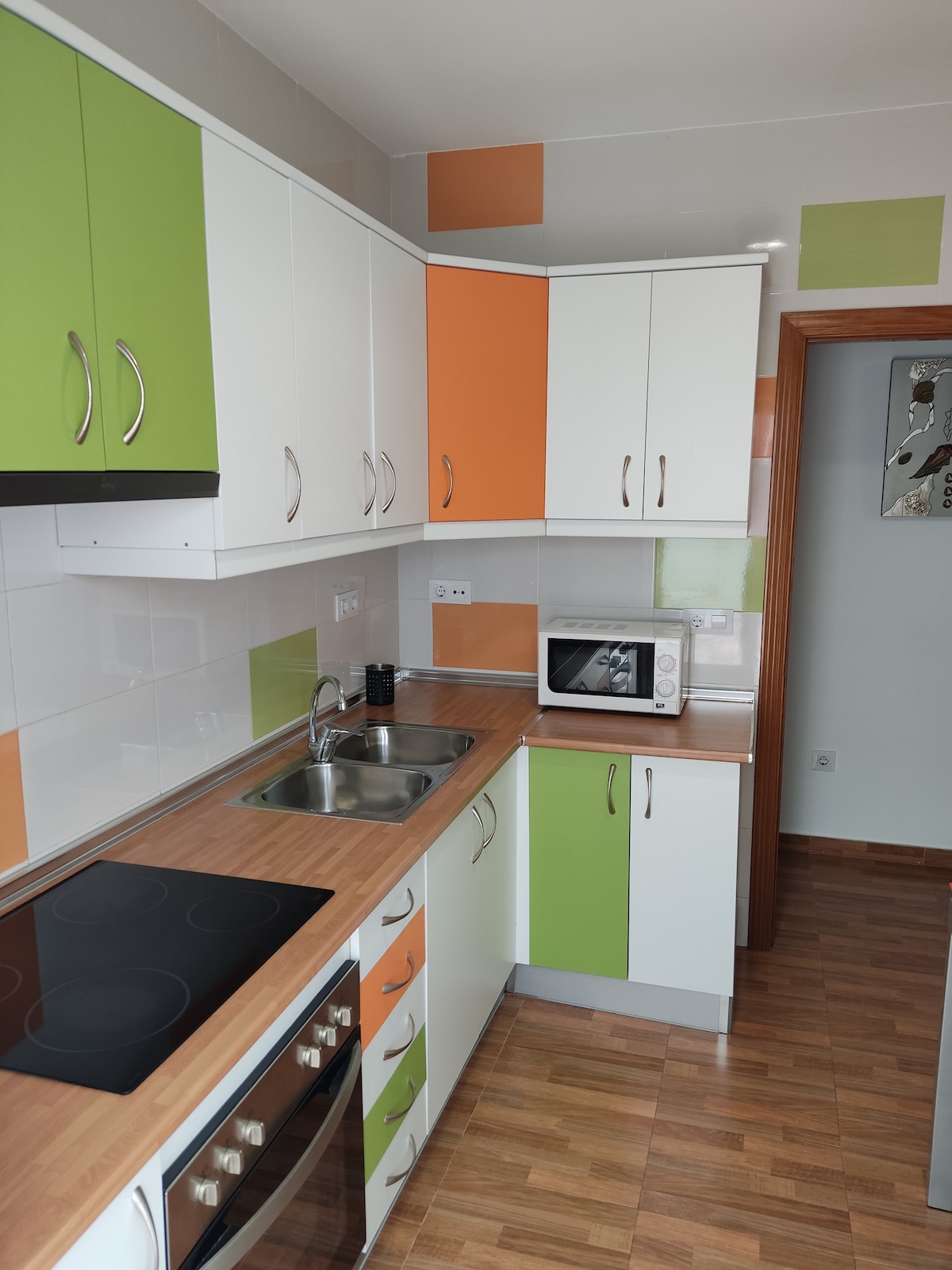 The kitchen features a contemporary design with white cabinets accented by vibrant green and orange panels. A stainless-steel sink is positioned beneath a countertop, with an electric cooktop and oven below. A microwave is located on the counter, enhancing functionality.