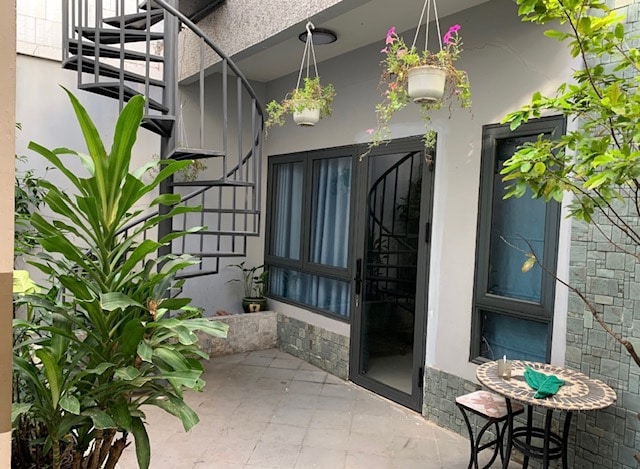A spiral staircase is seen leading to the upper level, surrounded by green plants. The entryway features a glass door adjacent to large windows. A small table with chairs is positioned on the patio, providing a serene outdoor space with a secluded atmosphere.