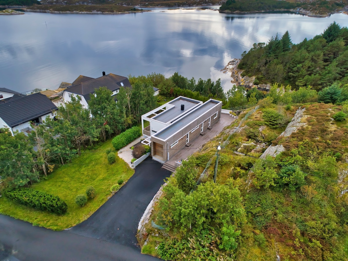 A modern cabin is set in a tranquil neighborhood, surrounded by greenery and rocky outcrops. The flat-roofed design and expansive windows provide views of the calm water and nearby islands, showcasing the integration of nature and architecture.