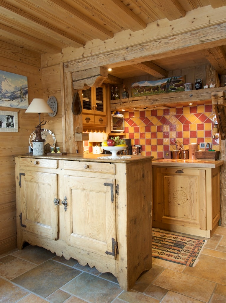 A rustic wooden sideboard is positioned against a wall, showcasing a collection of decorative items and a lamp. Adjacent to it, a compact kitchen area features colorful tiled backsplash and wooden cabinetry, creating a warm, inviting cooking space.