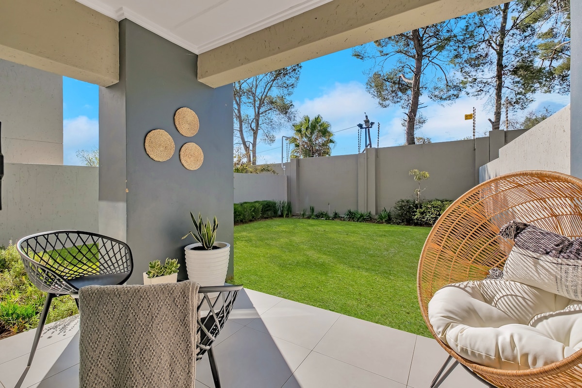 A private patio area features two outdoor chairs positioned around a small table. Lush greenery and well-maintained grass can be seen beyond the patio, while modern wall decor and a potted plant add visual interest to the serene outdoor space.