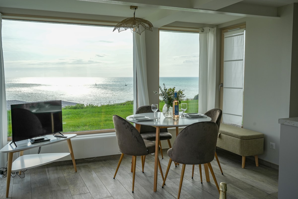 A dining area features a round table with four elegant chairs, positioned near large windows that provide panoramic views of the sea. Natural light floods the space, highlighting the coastal scenery and soft furnishings. A small television is placed nearby on a minimalist stand.