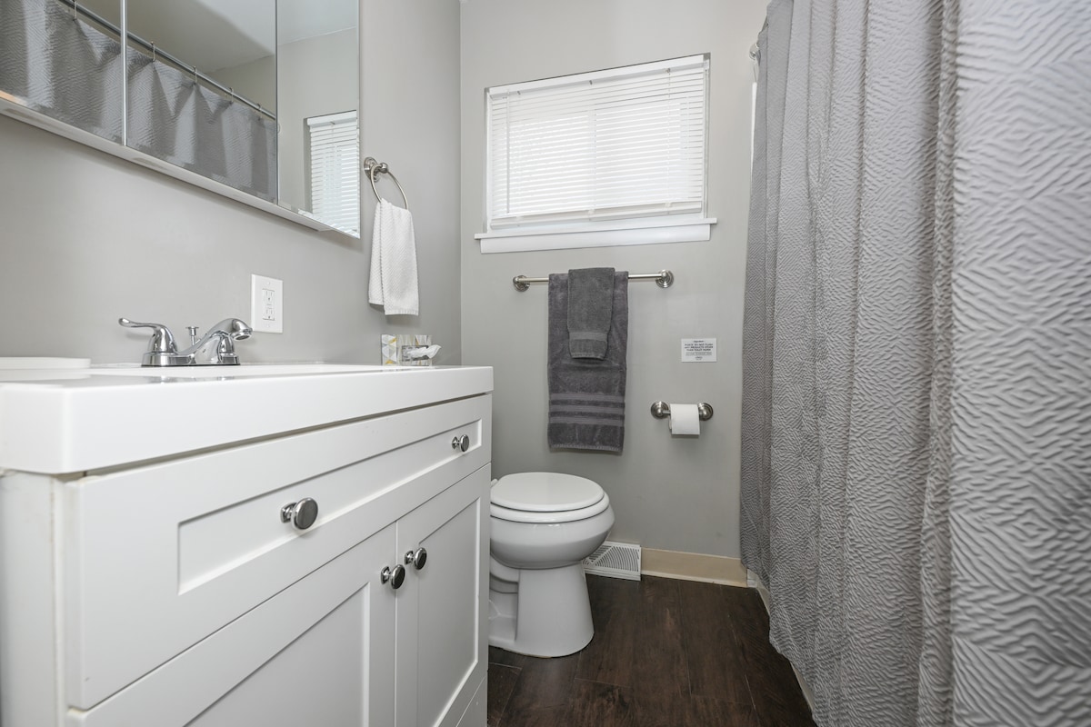A clean bathroom space features a white vanity with storage drawers and a large mirror above. A modern toilet is positioned next to a gray shower curtain. Bath towels are hung neatly on a rack, and natural light filters in through a window covered by sheer blinds.