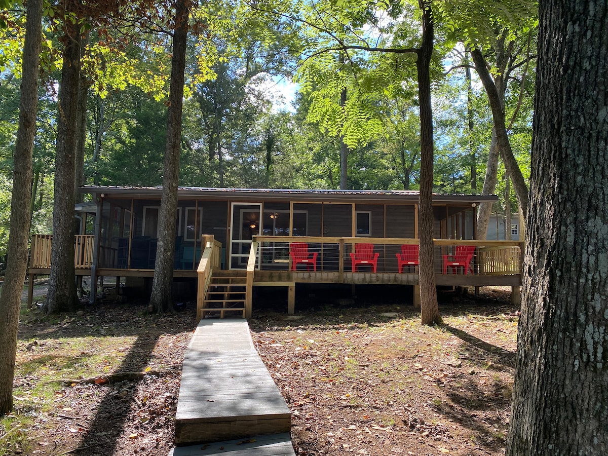 A charming two-bedroom house is set amidst trees, featuring a large wooden deck with several red chairs. A front stairway leads to the entrance, offering a welcoming path through the natural surroundings.