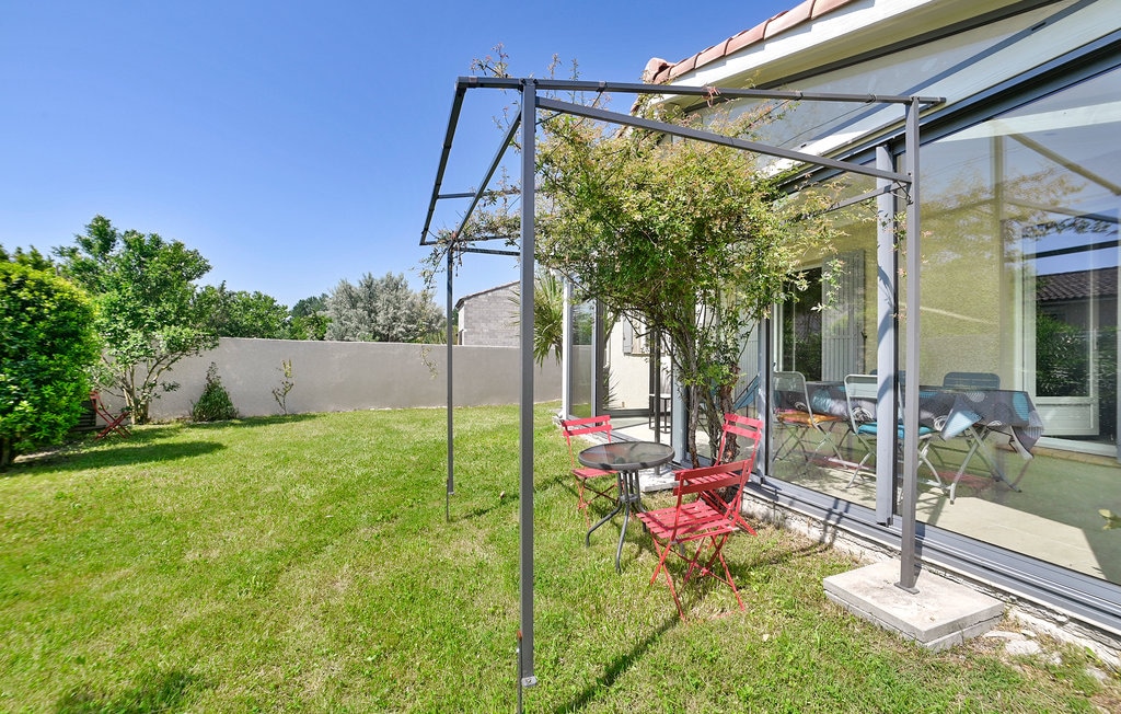 A spacious garden area is framed by a metal pergola, providing shade to a small table and red chairs. Lush green grass surrounds the space, with trees and shrubs adding greenery to the landscape. Large glass doors are visible, connecting the interior to the outdoors.