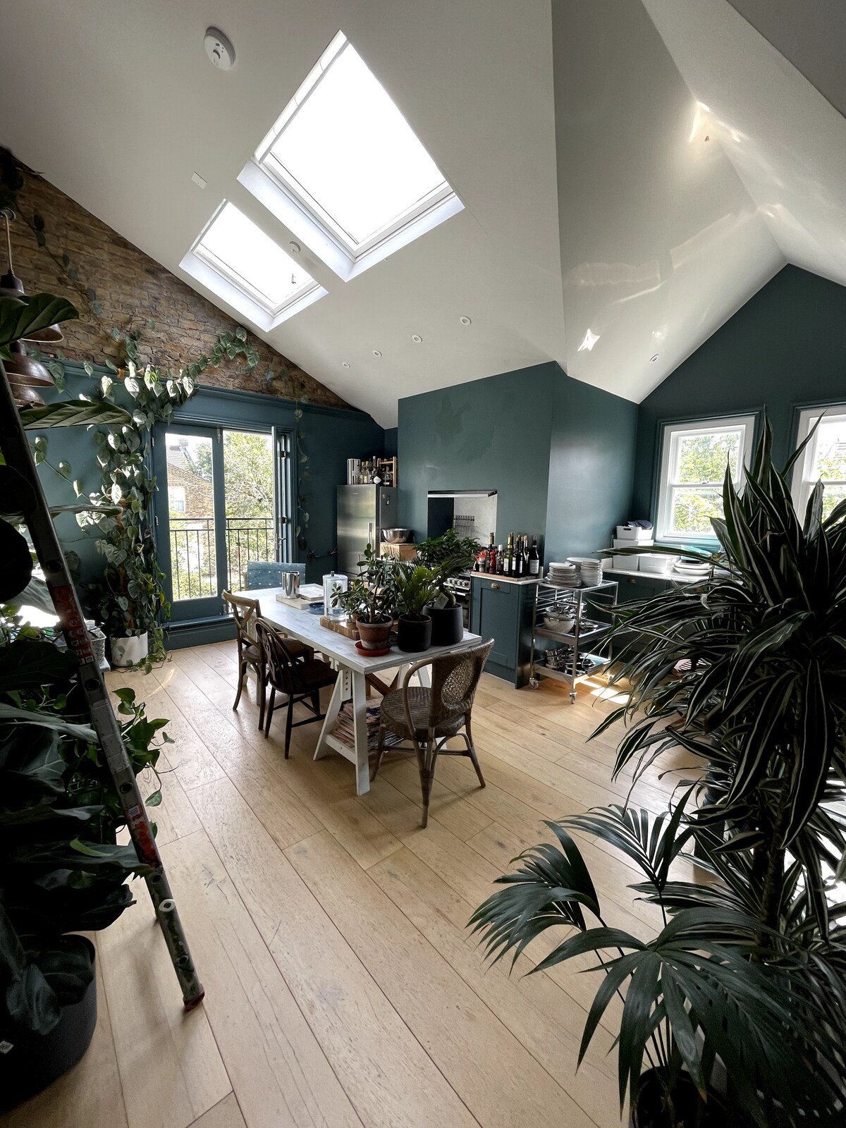A spacious kitchen and dining area features high ceilings with skylights that allow natural light to fill the space. A long wooden dining table is surrounded by chairs, and various indoor plants add a vibrant touch to the room. Large windows offer views of the outside.