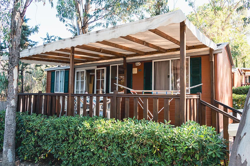 A charming bungalow is shown with a covered porch, surrounded by lush greenery. Wooden railings encase the spacious outdoor area, while large windows allow natural light to enter. The roof provides shade, creating a comfortable retreat for relaxation.