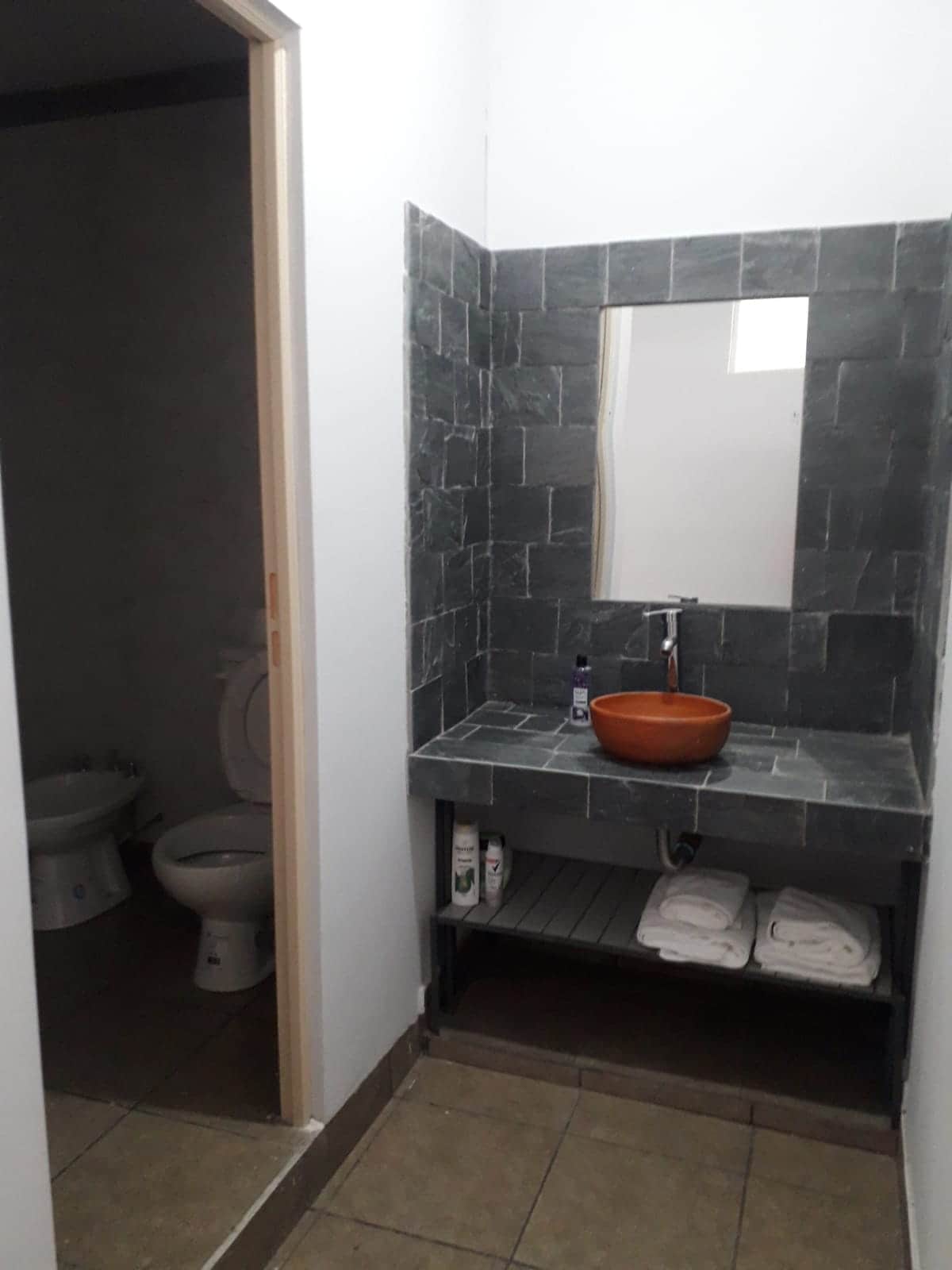A modern bathroom is highlighted by a sleek stone sink and wooden bowl. The wall features dark stone tiles, while light tones create a balanced atmosphere. Shelves hold neatly arranged towels and toiletries. A separate area is visible for the toilet.