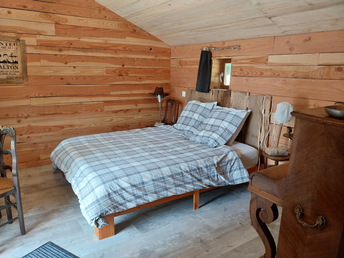 A cozy bedroom features wooden paneling and a spacious bed with a checked blanket. Two wooden chairs are positioned near the bed, and a vintage dresser is visible, enhancing the rustic charm of the space.